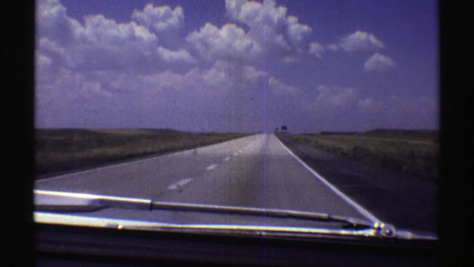 Late-1960s 8mm view from a car on a Nebraska highway, with flat plains and large clouds filling the upper frame.