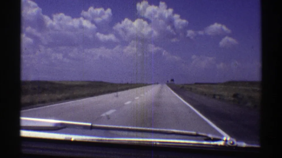 1969 Nebraska 8mm frame showing a car-dashboard view down a long, open highway with plains and large clouds ahead.