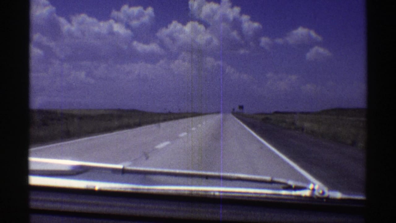 1969 Nebraska road-travel frame with the dashboard and wiper visible as the highway runs through open plains under a blue-purple sky.
