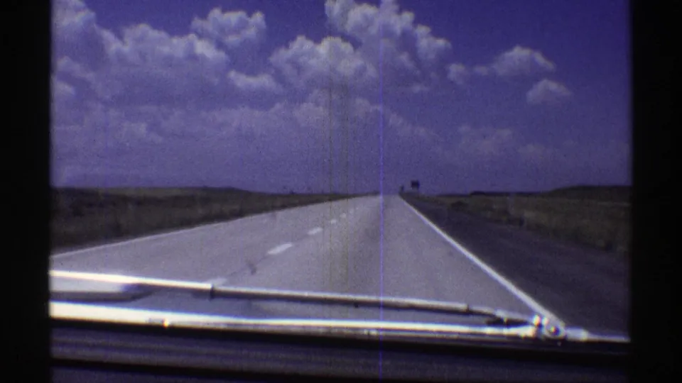 1969 Nebraska road-travel frame with the dashboard and wiper visible as the highway runs through open plains under a blue-purple sky.