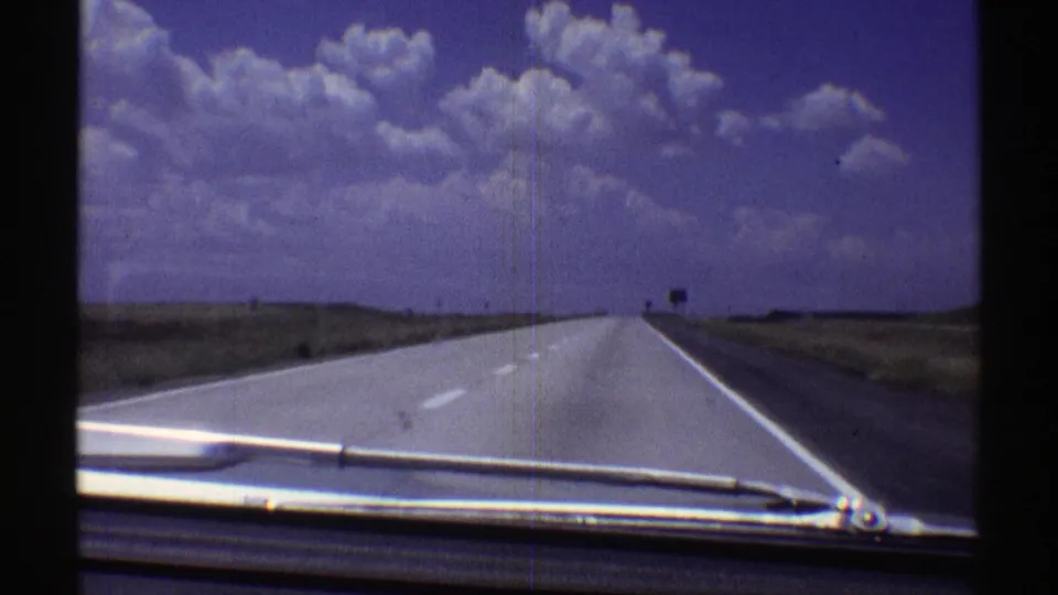 Late-1960s Nebraska home-movie frame of a long rural highway viewed through a car windshield under a broad clouded sky.