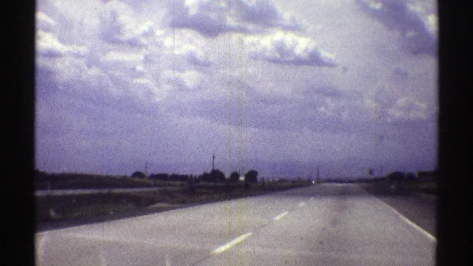A 1969 Nebraska road scene shows a broad highway, low horizon, sparse roadside features, and mottled cloud cover on 8mm film.