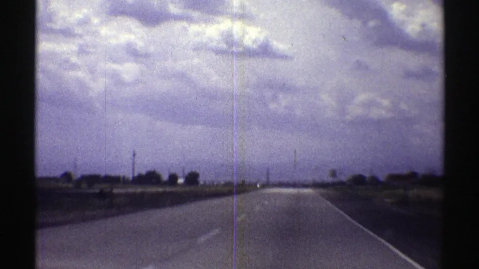 The 8mm frame shows a Nebraska highway receding into the distance, bordered by open ground and a sparse line of trees or structures.