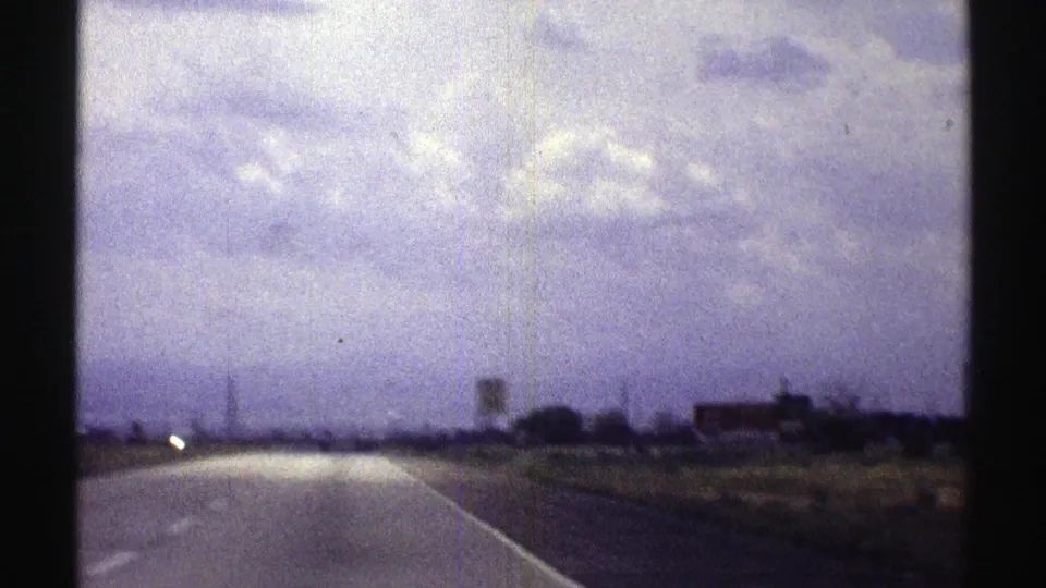 A late-1960s Nebraska highway view appears on faded 8mm film, with open roadway, low fields, distant structures, and clouded sky.