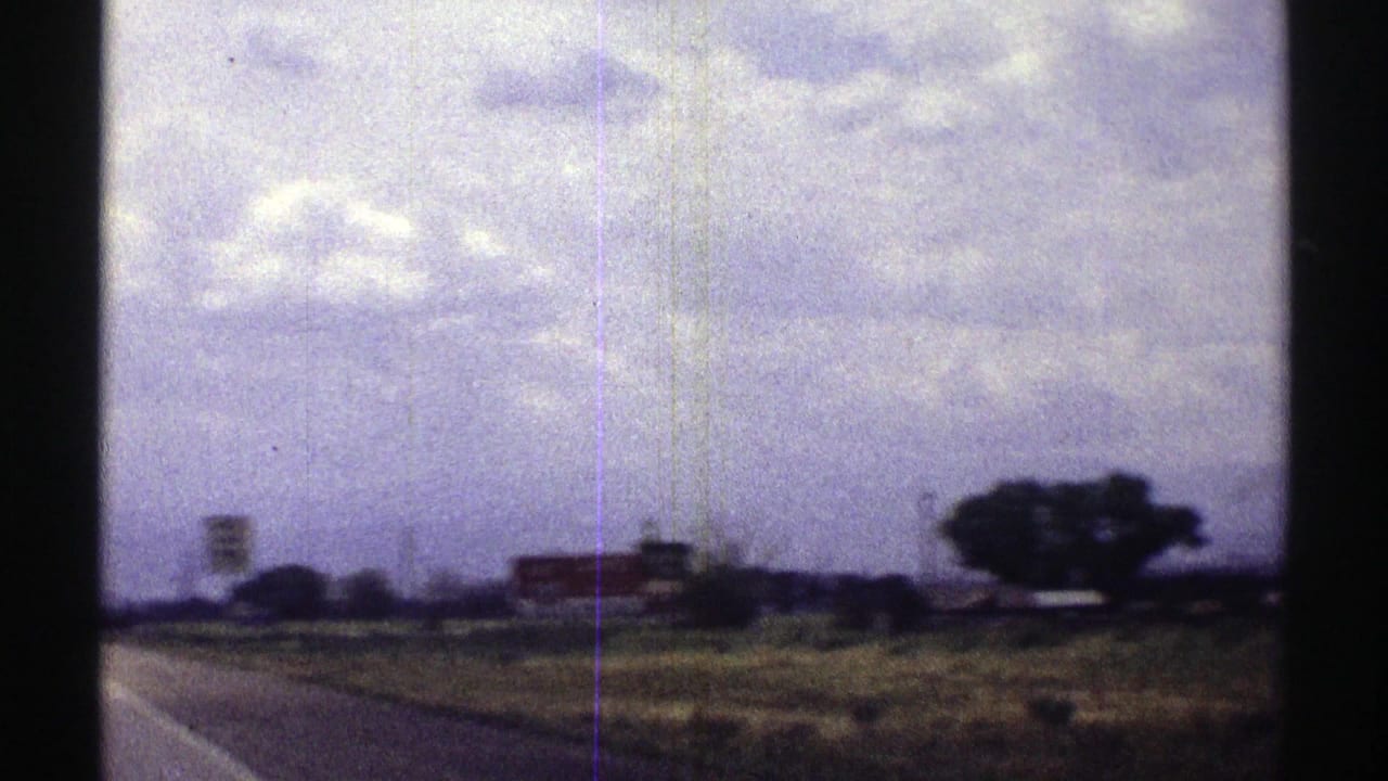 Rural highway shoulder with open fields, distant signs, trees, and a cloudy sky.