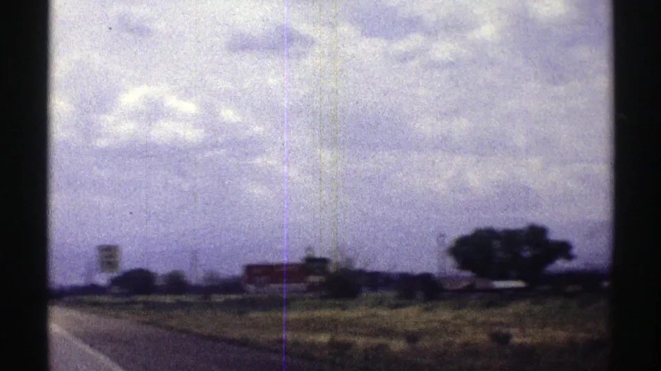 1969 Nebraska road-trip frame showing a rural highway edge, open fields, distant roadside signs, trees, and clouds.