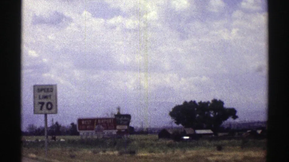 1969 Nebraska highway frame with a visible speed limit 70 sign, red roadside sign, open grass, trees, and cloud cover.