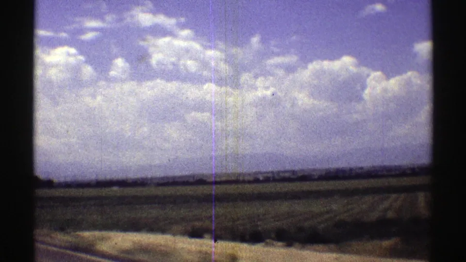 1969 Nebraska travel view of open rural fields, distant horizon, road edge, and a bright cloudy sky from a moving vehicle.