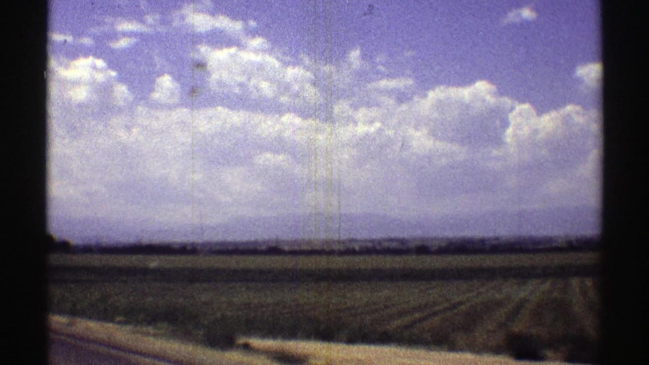 1969 Nebraska 8mm home-movie frame showing rural farmland rows stretching toward a low horizon under bright cumulus clouds.