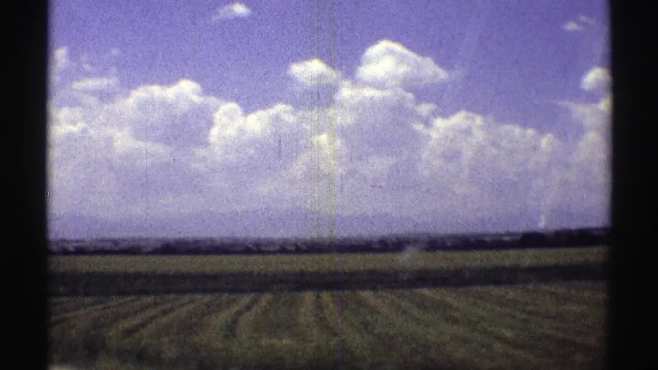 Vintage 8mm frame of Nebraska farmland from a moving road-view perspective, with soft grain and bright clouds above the horizon.