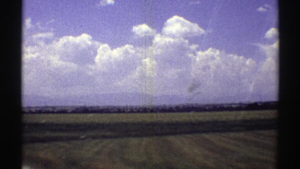 Late-1960s Nebraska home-movie frame showing open agricultural land and a wide sky with visible 8mm grain and scratches.