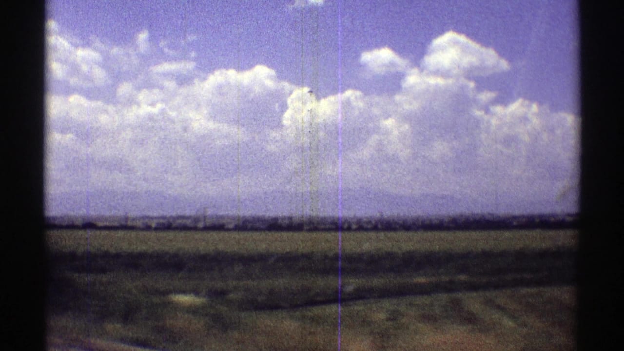 1969 Nebraska 8mm frame showing flat fields, a low horizon, and bright clouds from a moving road-trip viewpoint.