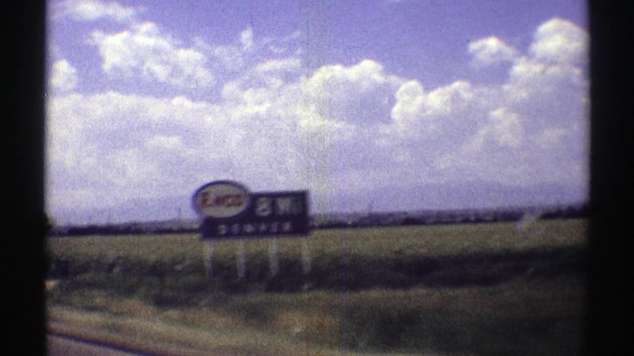 A roadside Enco sign passes beside open fields under a sunny sky.