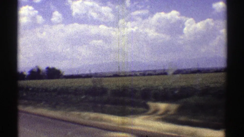 1969 Nebraska 8mm frame showing a roadside curve, flat fields, and a distant tree line under bright clouds.