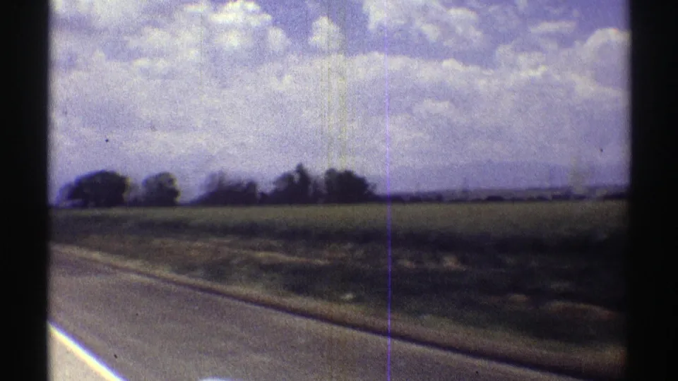 1969 Nebraska 8mm frame with paved roadside, open field, distant trees, and a bright clouded sky.