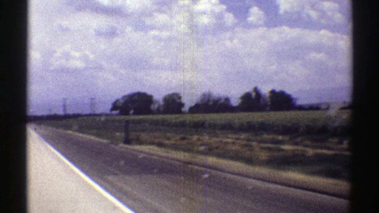 1969 Nebraska 8mm view from a moving vehicle along a rural highway bordered by fields, trees, and heavy clouds.