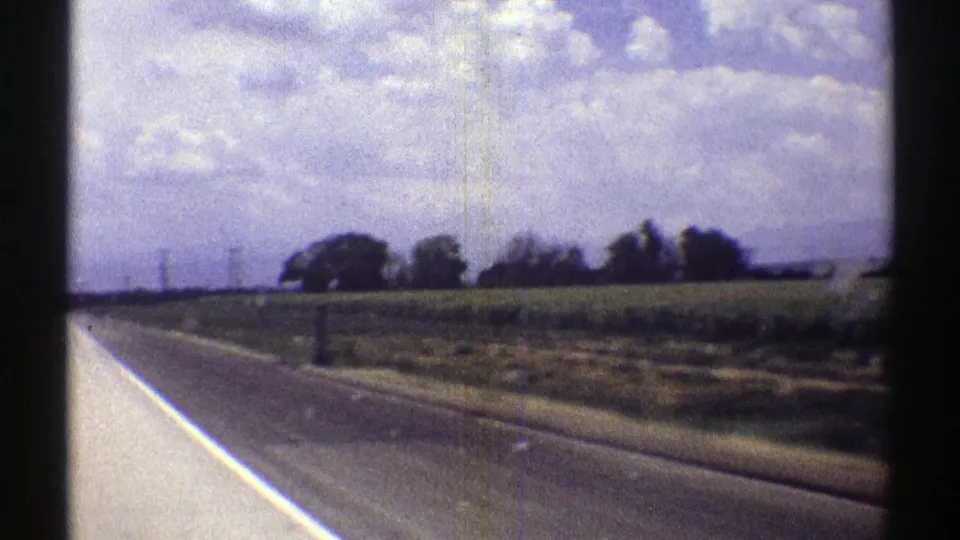 1969 Nebraska 8mm view from a moving vehicle along a rural highway bordered by fields, trees, and heavy clouds.