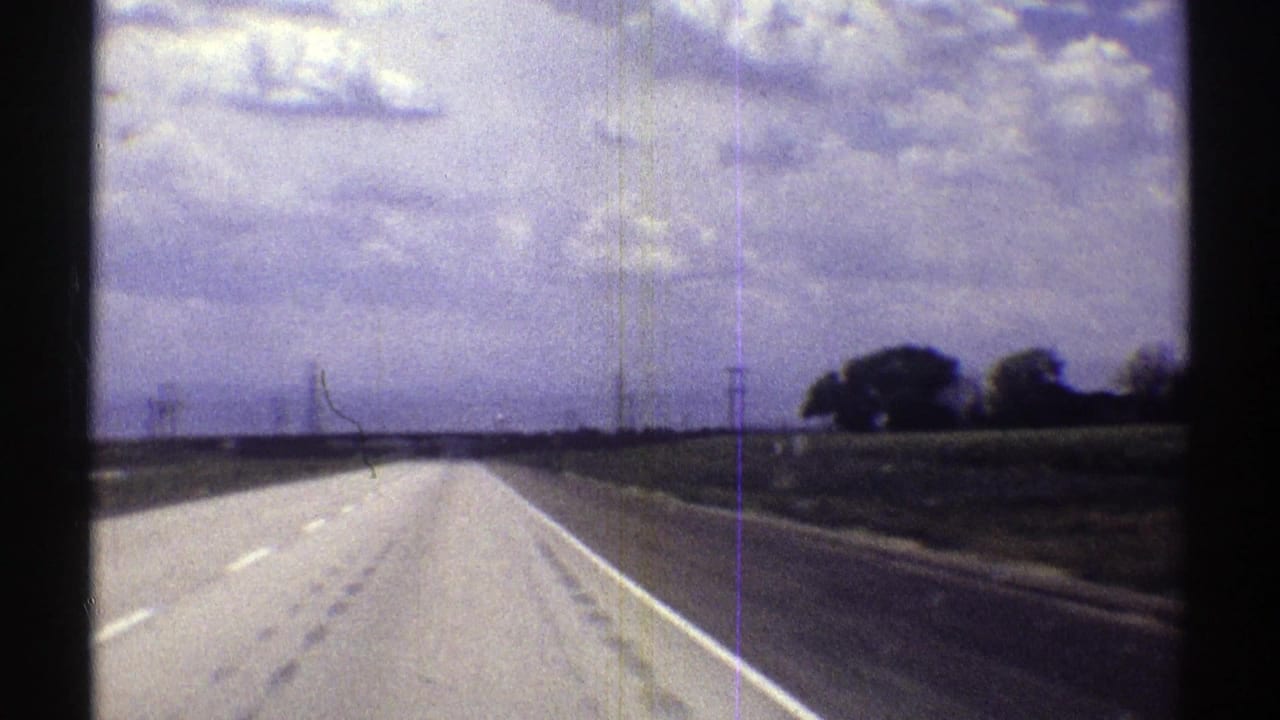 Forward-looking 1969 Nebraska road view showing a straight highway, roadside fields, power lines, and dense cloud cover.
