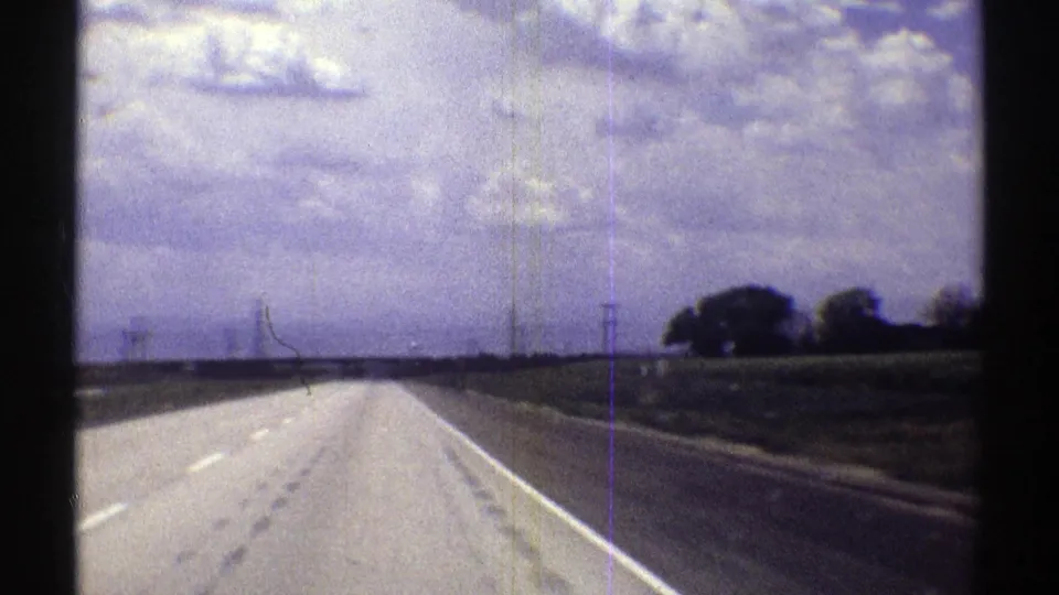 Forward-looking 1969 Nebraska road view showing a straight highway, roadside fields, power lines, and dense cloud cover.
