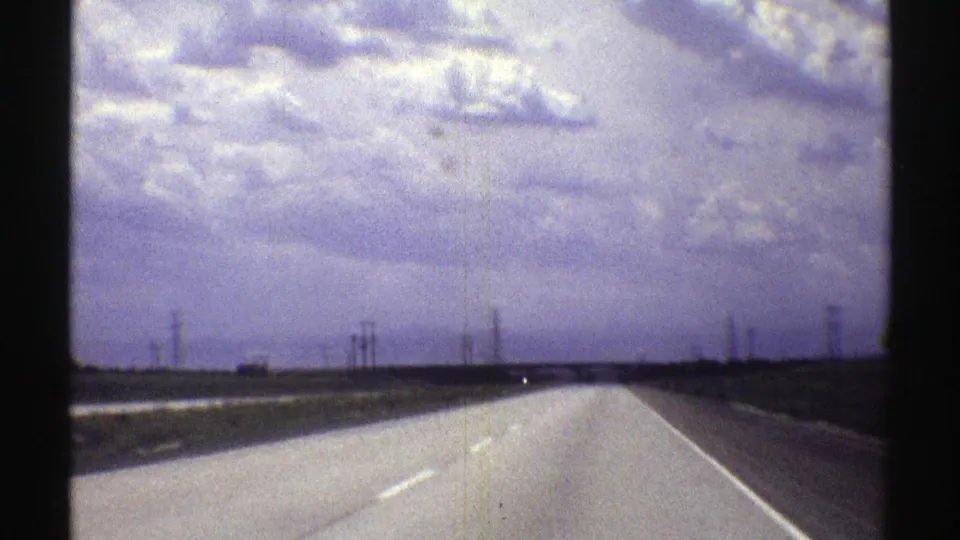 Vintage 8mm Nebraska highway scene with a low horizon, distant transmission towers, open land, and a heavy overcast sky.