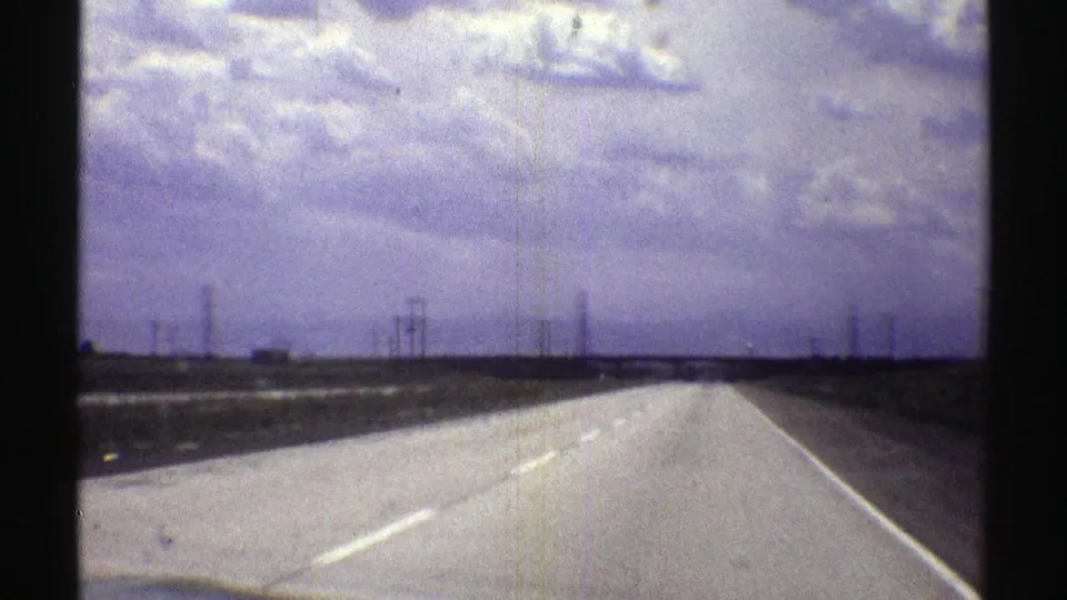 1969 Nebraska 8mm driving view with highway pavement, lane markings, fields, utility towers, and gray storm clouds.