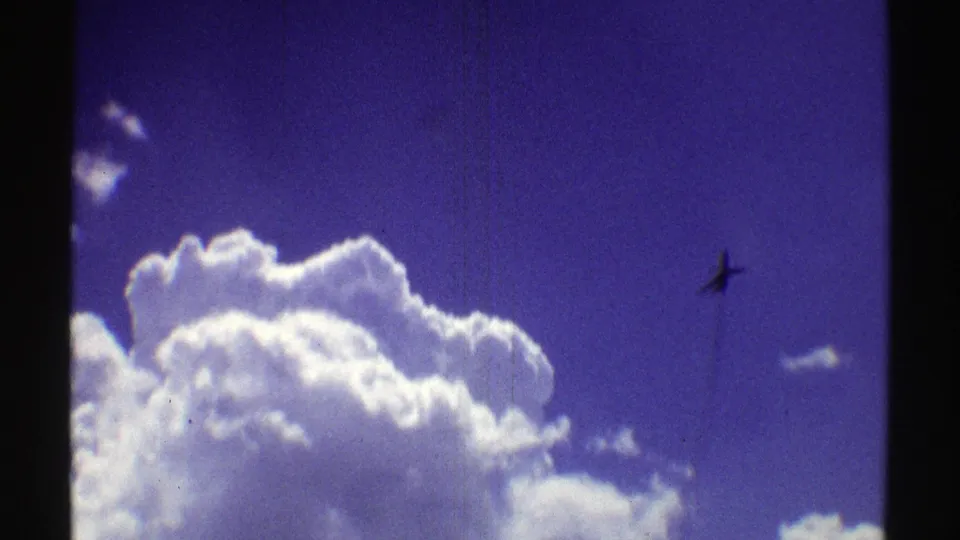 1969 Nebraska home-movie frame with sunlit cumulus clouds and a small airplane silhouetted in blue sky.