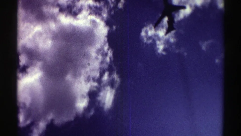 1969 Nebraska home-movie frame with a jet-like airplane silhouette crossing near sunlit clouds.