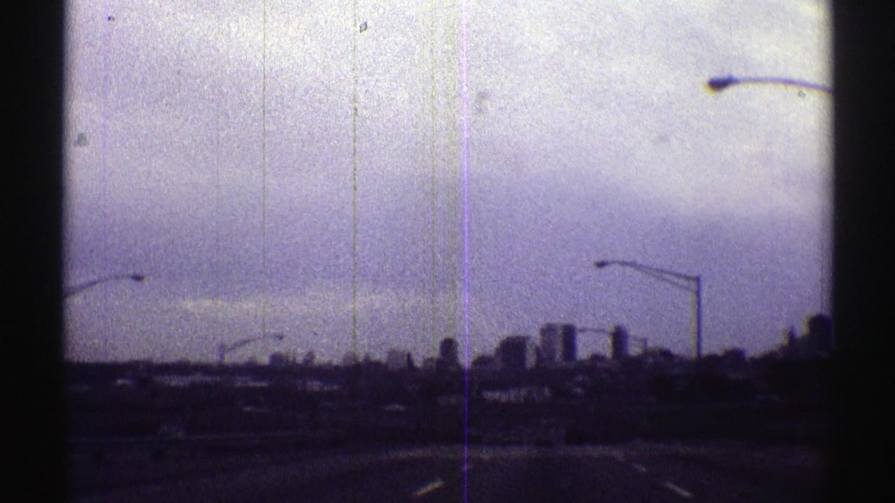 Distant Nebraska city skyline appears ahead from a moving roadway, with streetlight poles silhouetted below a pale sky.