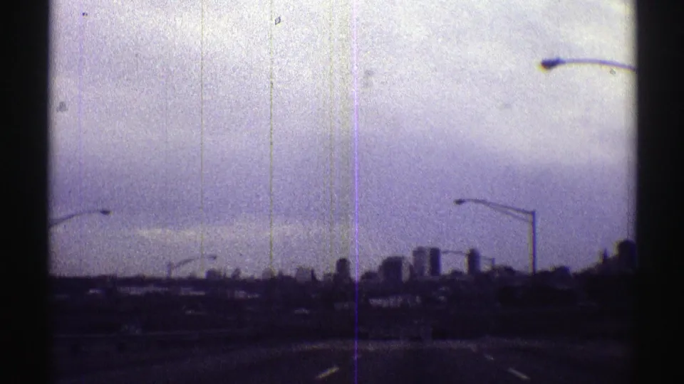 Distant Nebraska city skyline appears ahead from a moving roadway, with streetlight poles silhouetted below a pale sky.