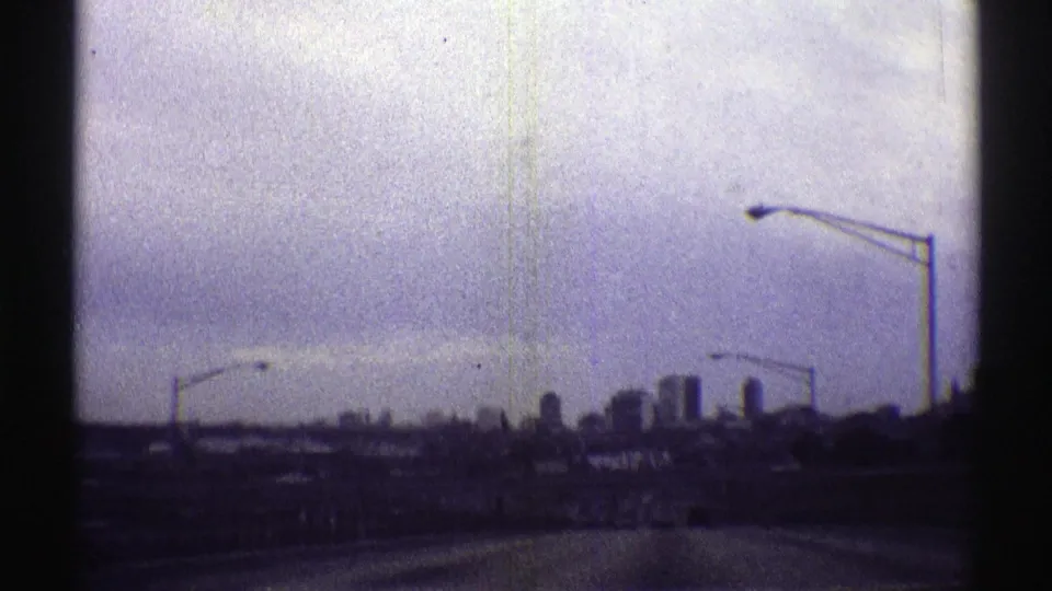 Moving road view toward a distant Nebraska skyline, with multiple streetlight poles and low urban silhouettes under a gray-blue sky.