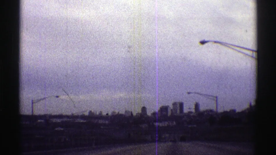 Distant city buildings and roadside streetlights remain in view from the moving Nebraska road approach, with purple-gray 8mm wear visible.
