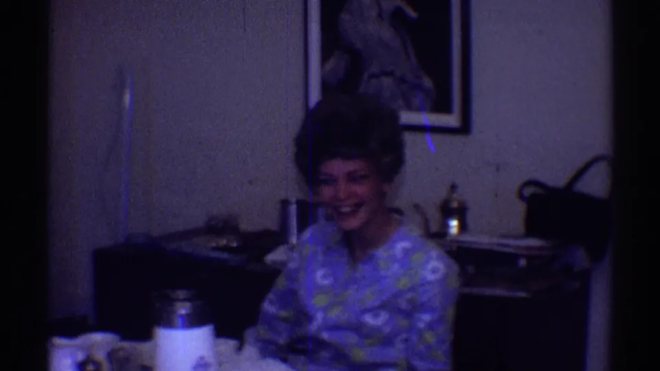 In a 1969 Clarendon Hills home-movie frame, an adult woman in a floral dress smiles while seated at a table near a white thermos.