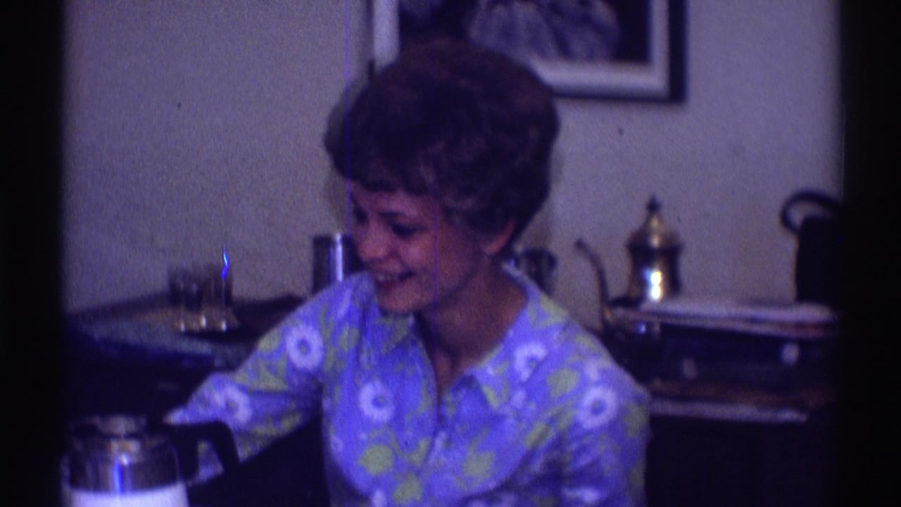 Short-haired woman in a blue floral shirt sits at a table, smiling toward someone off camera.