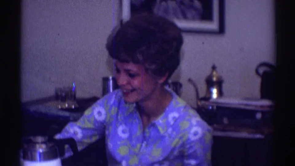 In a 1969 Clarendon Hills home interior, a short-haired woman in a blue floral shirt smiles while seated at a dining table.