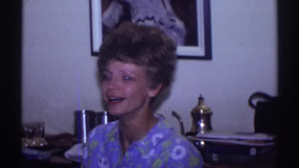 The 1969 home-movie frame shows the short-haired woman smiling and speaking near a dining table in a private Clarendon Hills interior.