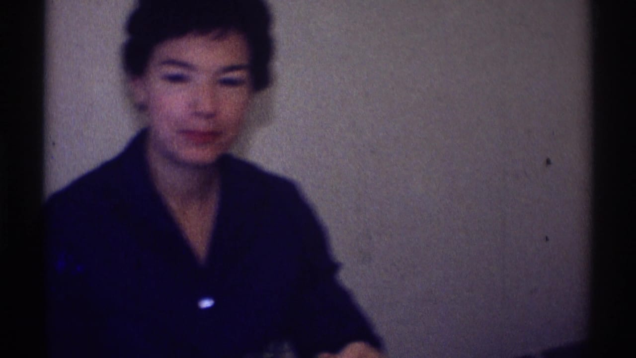 Woman in a dark collared top seated against a plain wall in a home interior.