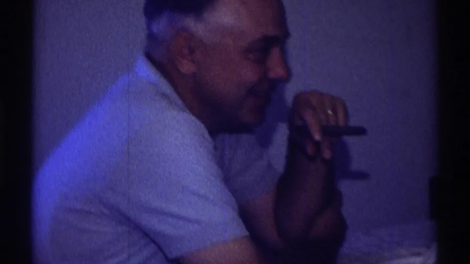 A man in a light shirt sits in profile inside the private home setting, holding a cigar in the 1969 Clarendon Hills footage.