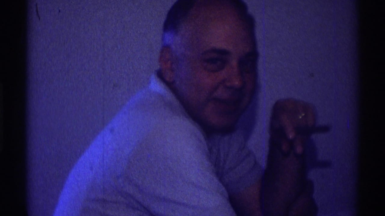 In a 1969 Clarendon Hills home-movie frame, a seated man in a light polo shirt looks toward the camera while holding a cigarette.