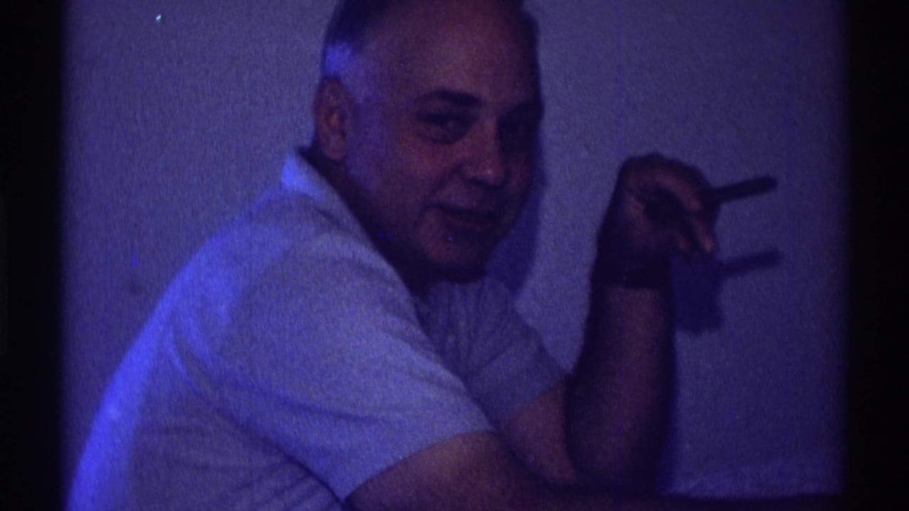 A seated adult man in a light shirt smiles faintly toward the camera, cigarette raised, in a blue-lit private interior.