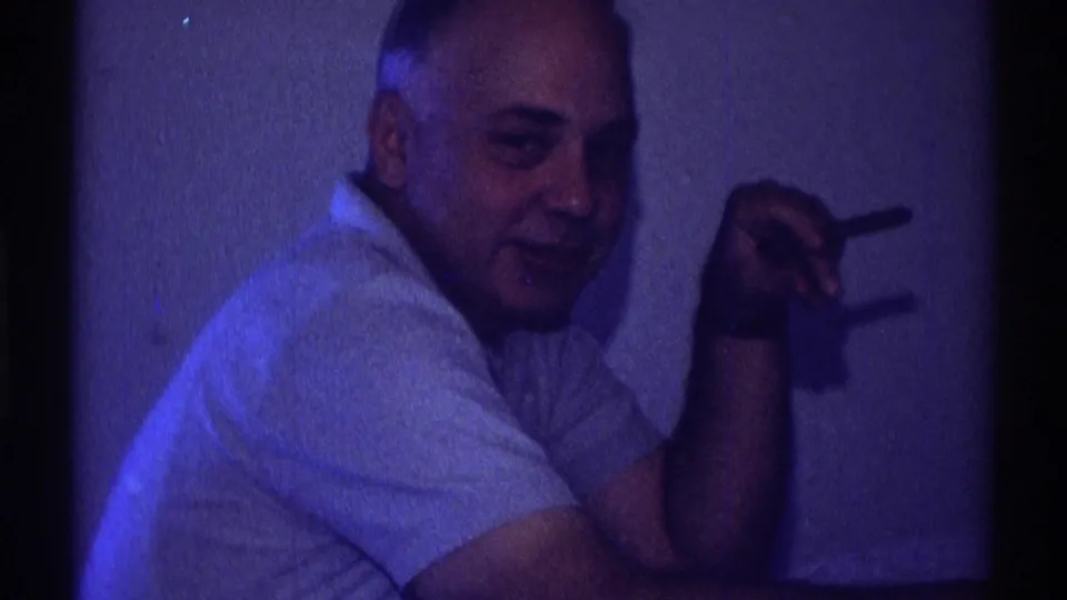 A seated adult man in a light shirt smiles faintly toward the camera, cigarette raised, in a blue-lit private interior.