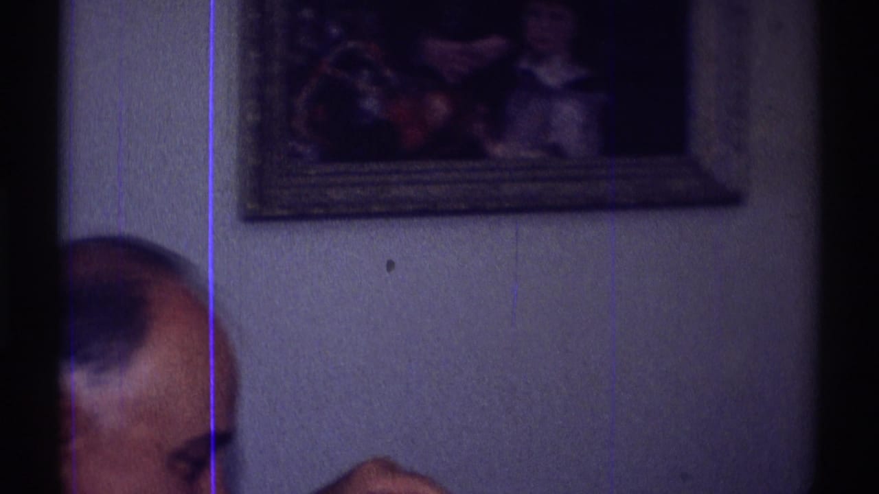 Cropped bald man at left with framed wall picture behind him in a dim interior.