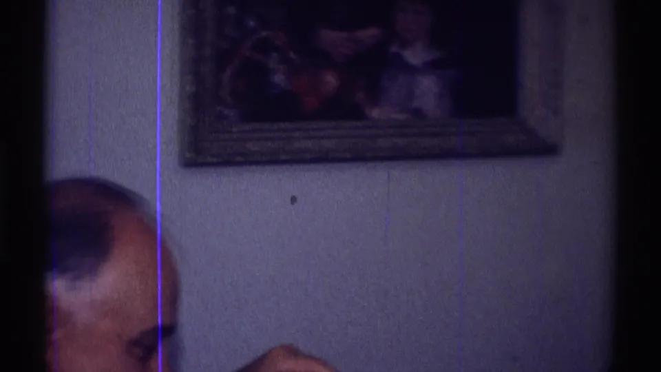 In a 1969 Clarendon Hills home-movie frame, a cropped seated man appears beside a wall with framed artwork.