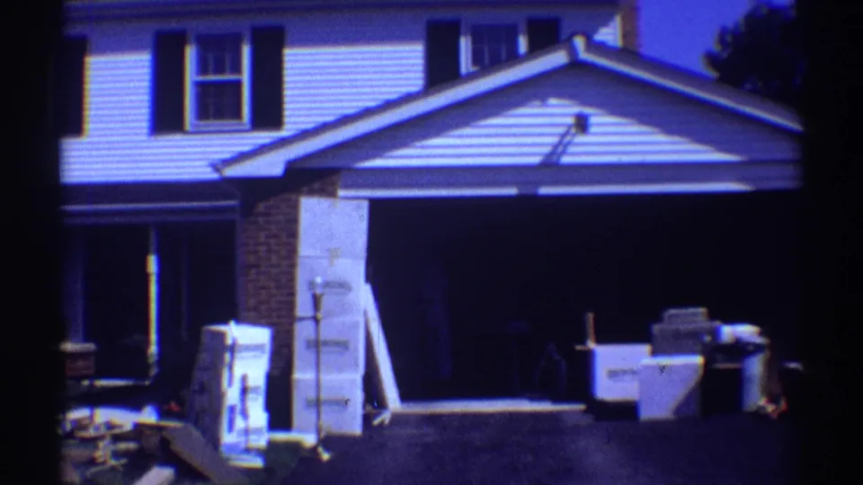 1969 Clarendon Hills home-movie frame showing a white suburban house with dark shutters and moving boxes or household goods outside.