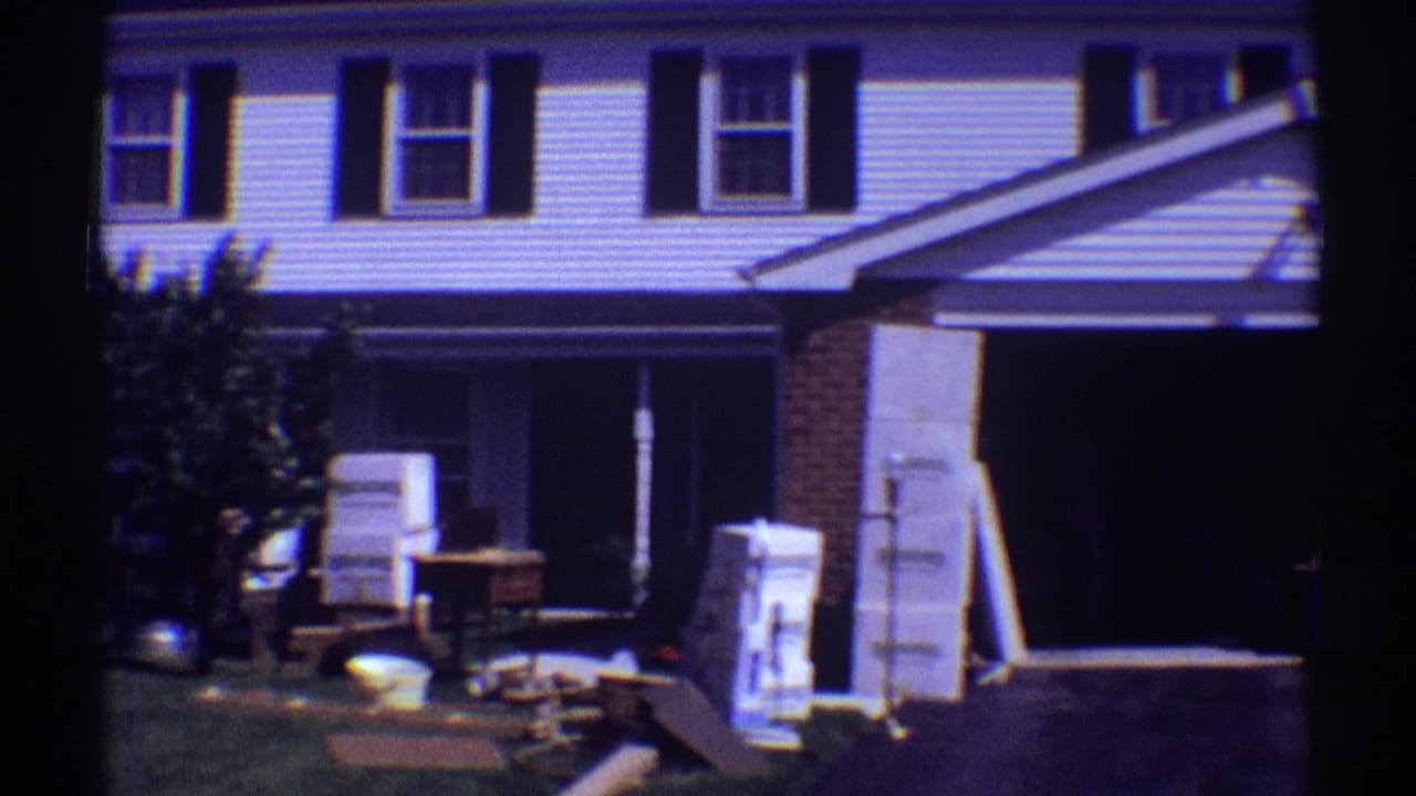 1969 Clarendon Hills home-movie frame showing a white two-story house with dark shutters, lawn items, boxes, and furniture during a move.