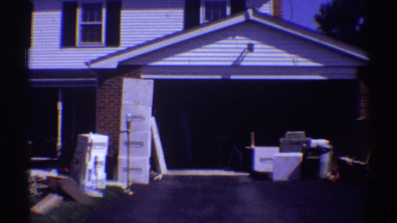 Open garage beside a driveway with boxes, furniture, and household items outside.