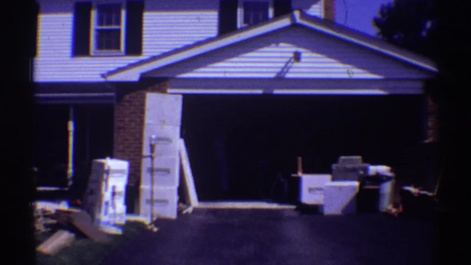 1969 Clarendon Hills frame centered on an open garage, driveway, stacked boxes, and furniture set outside a white suburban house.