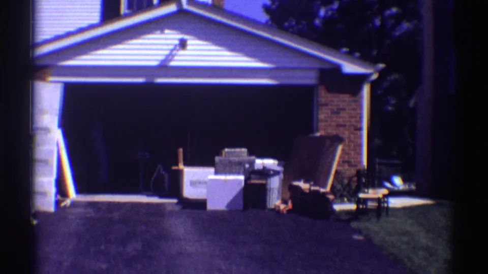 1969 Clarendon Hills home-movie frame showing the open garage, driveway, boxes, furniture, and yard edge during a residential move.