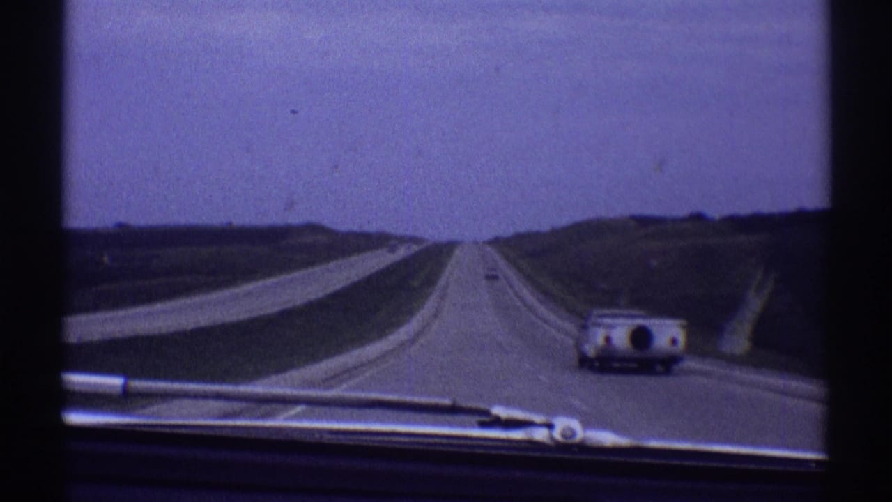 Straight highway seen through a windshield with a camper-towing car ahead on the right.