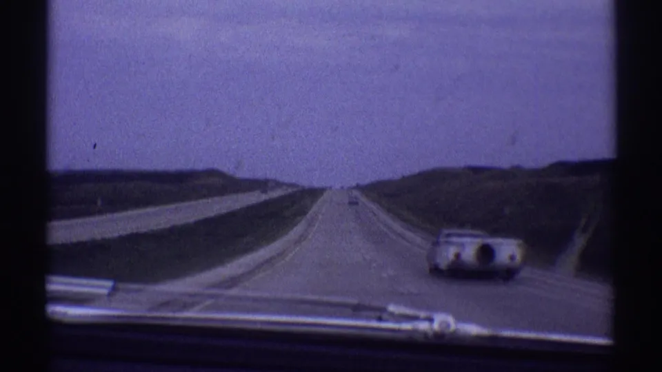 Vehicle-mounted 8mm view of a 1969 highway scene, with a rounded camper trailer car visible to the right and open road ahead.