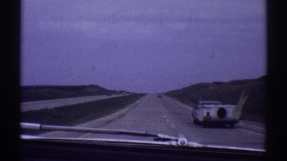 Interior car perspective on a 1969 Illinois highway, showing windshield hardware and a camper-towing vehicle traveling nearby.